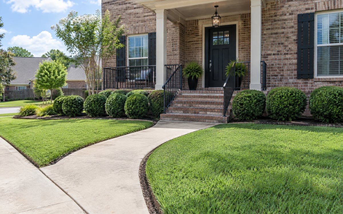 A concrete walk curves to the brick front steps of a tidy house, with trimmed boxwood balls in the beds and a dark green St. Augustine lawn flush to a clean edge.