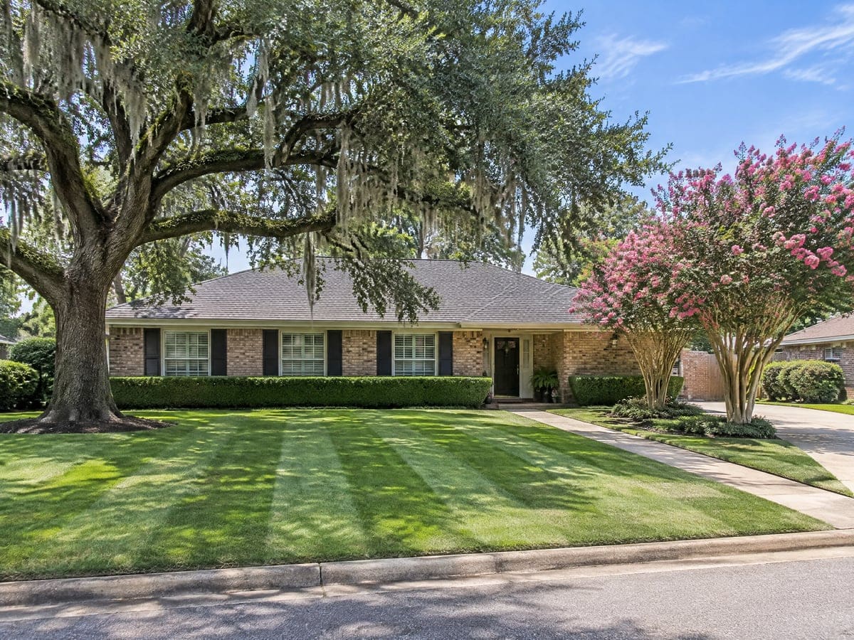 A one-story brick house under a sprawling live oak draped with Spanish moss, with a wide front lawn mowed in alternating lawn care stripes down to the street.