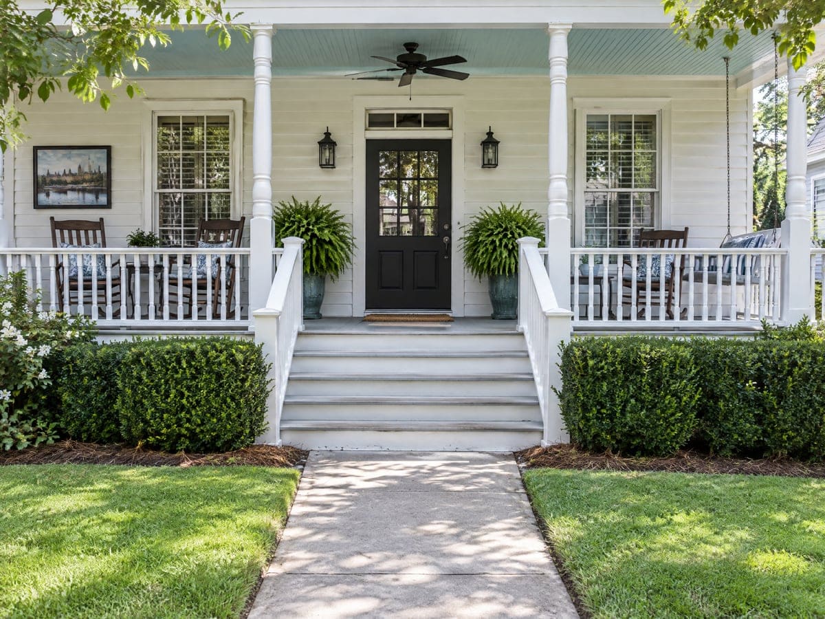 The quiet front porch of a white one-story house with rocking chairs on both sides of the door, hanging ferns and a dark front door above wide steps, a home that might call a lawn service.