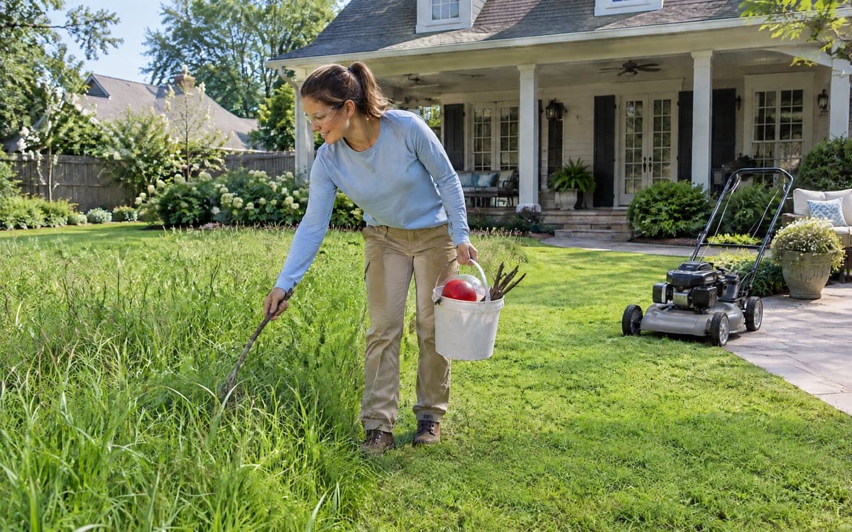 A woman in safety glasses walks a long patch of overgrown grass in the back yard with a pickup bucket in one hand, bending to reach for a fallen stick before mowing it down.