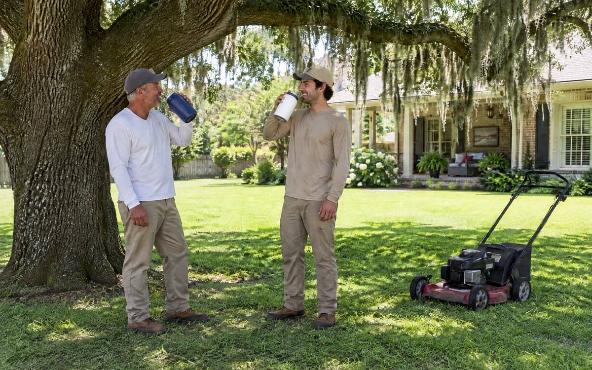 Two men take a water break under a wide live oak draped with Spanish moss, with a red push mower parked on the grass behind them and a white cottage's front porch at the far right.