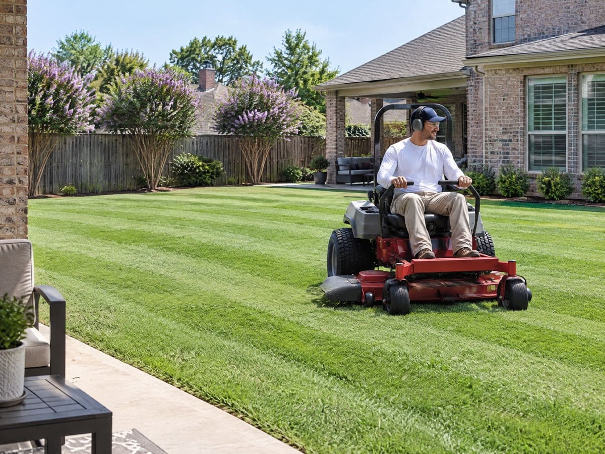 A worker on a red zero-turn mower lays alternating stripes into a thick back lawn beside a brick house on a weekly lawn visit, with crape myrtles in bloom along the back fence.